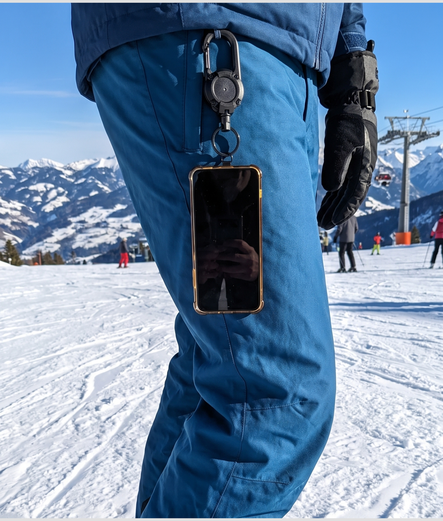 Telefoon aan een uitrolbaar koord aan een blauwe skibroek, met de piste en een stoeltjeslift op de achtergrond